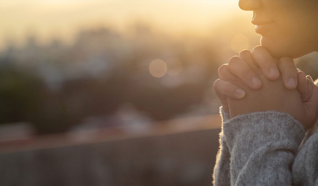 Woman in sweater praying at sunrise with city in the background.