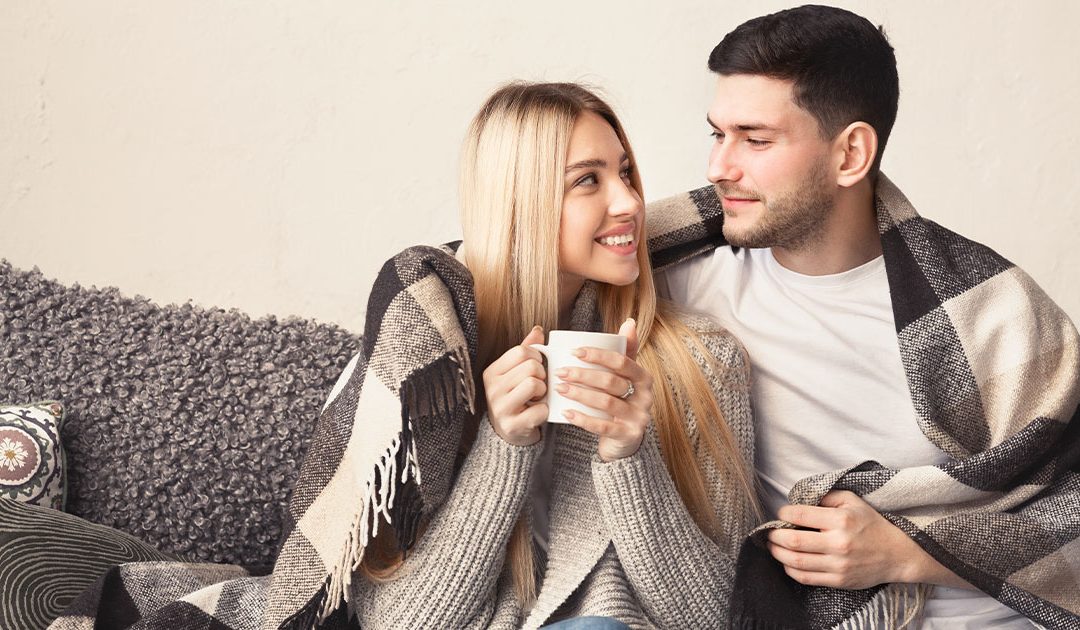 A couple sits and shares a blanket; the woman enjoys coffee, and they look at each other, smiling.